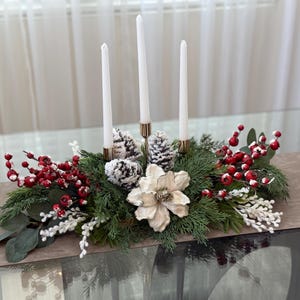 May include: A festive centerpiece featuring three white candles in gold holders, surrounded by greenery, red berries, white accents, and pinecones. A large, cream-colored flower adds a touch of elegance to this holiday decoration.