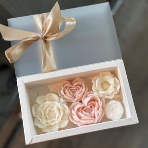 May include: A white gift box with a clear lid, tied with a gold satin ribbon. Inside, six floral-shaped soaps in shades of white and pink are arranged. The soaps include peony, rose, and heart-shaped designs.