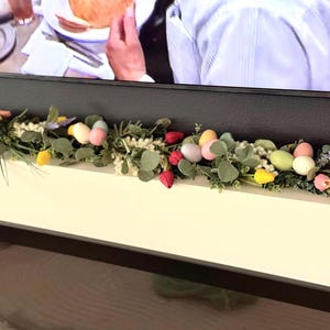 Może przedstawiać: Zielona girlanda z pastelowymi jajkami wielkanocnymi i czerwonymi tulipanami na białym kominku.