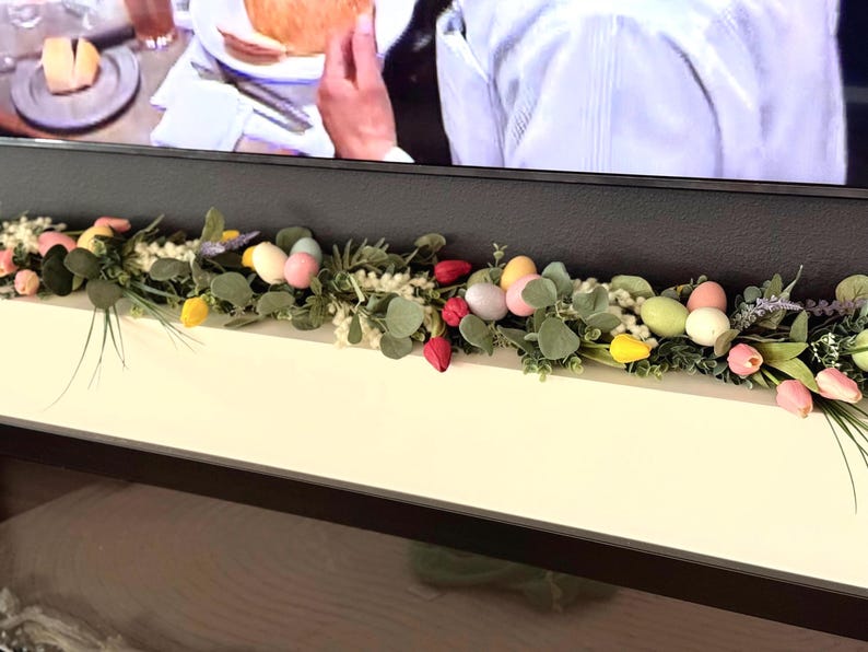 Może przedstawiać: Zielona girlanda z pastelowymi jajkami wielkanocnymi i czerwonymi tulipanami na białym kominku.