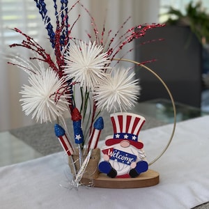 May include: A patriotic centerpiece featuring a gnome holding a sign that says "Welcome". The gnome is wearing a red, white, and blue hat and has a white beard. The centerpiece also includes white pom-poms, red and blue glitter sprigs, and silver firework-shaped decorations.