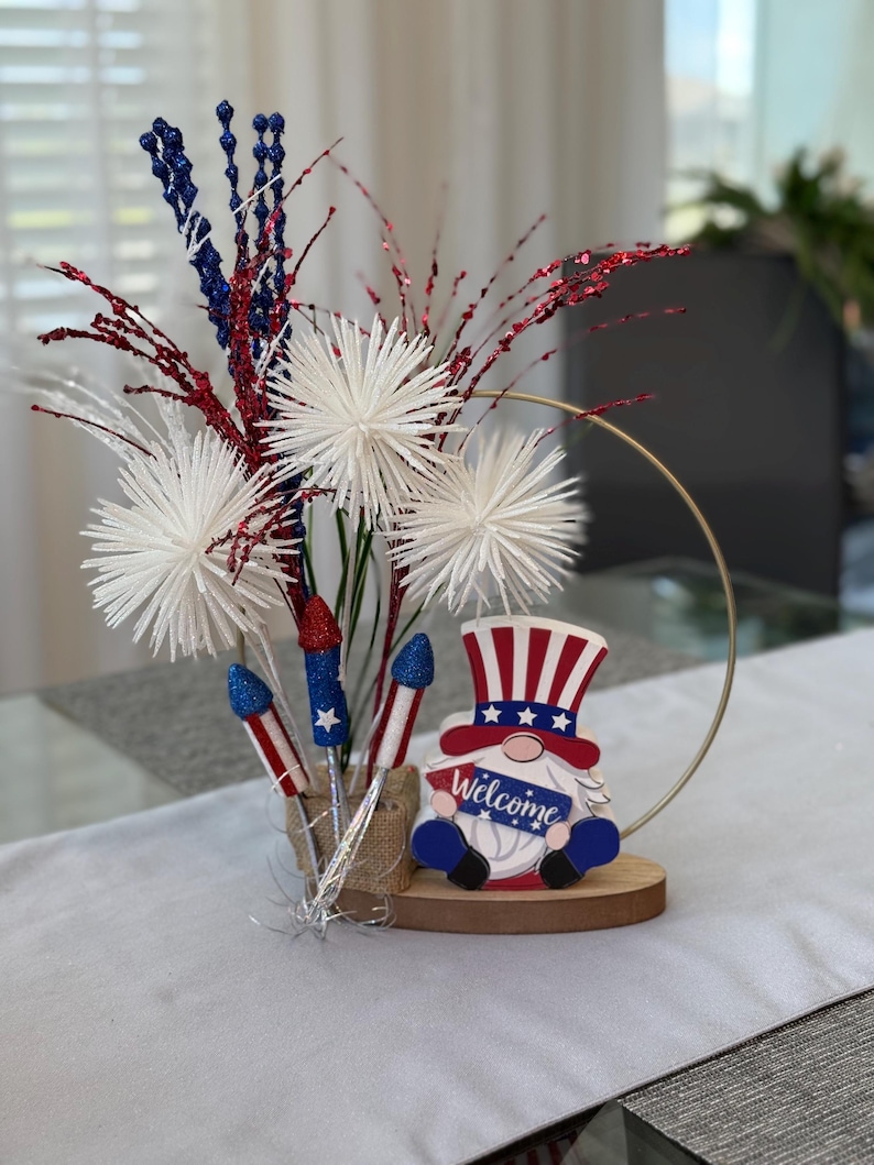 May include: A patriotic centerpiece featuring a gnome holding a sign that says "Welcome". The gnome is wearing a red, white, and blue hat and has a white beard. The centerpiece also includes white pom-poms, red and blue glitter sprigs, and silver firework-shaped decorations.