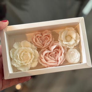 May include: A white box containing six floral-shaped soaps. The soaps are in shades of white and pink, with designs of roses and hearts. The box is held by a hand.