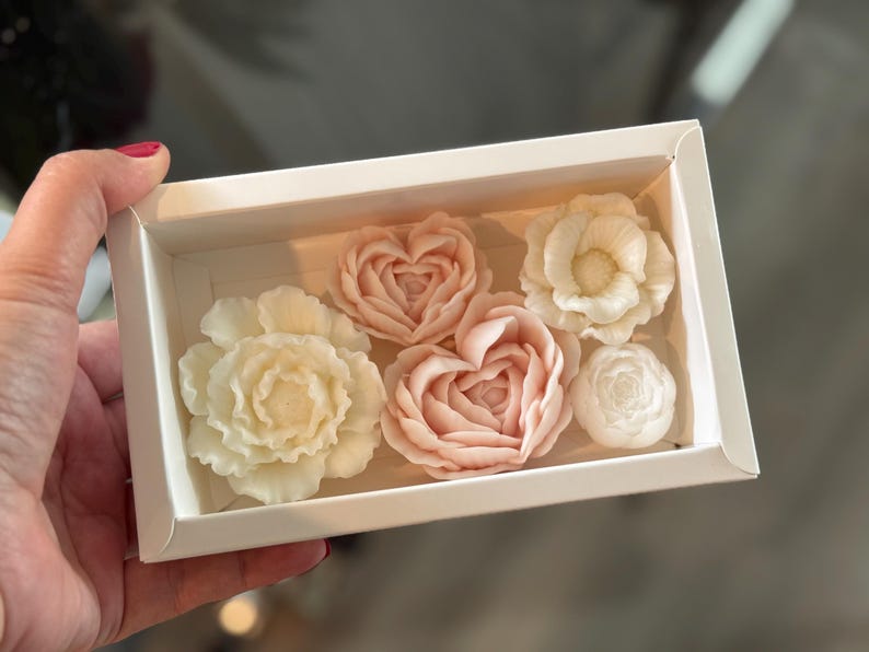 May include: A white box containing six floral-shaped soaps. The soaps are in shades of white and pink, with designs of roses and hearts. The box is held by a hand.