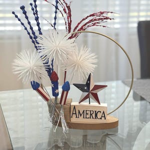 May include: A patriotic centerpiece featuring white pom-poms, red and blue glitter sprigs, and firework decorations. A wooden block with the word "America" sits on a wooden base with a gold hoop.