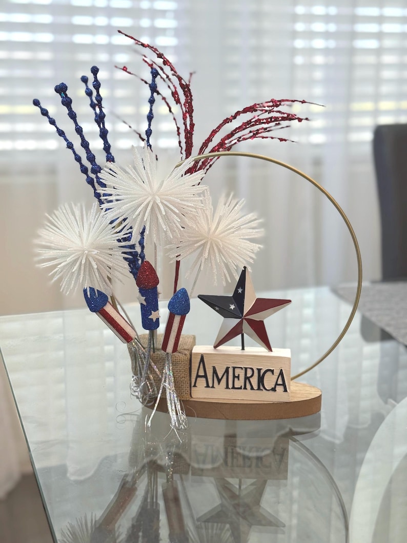 May include: A patriotic centerpiece featuring white pom-poms, red and blue glitter sprigs, and firework decorations. A wooden block with the word "America" sits on a wooden base with a gold hoop.