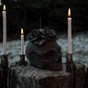 May include: A dark gray skull-shaped candle holder with a floral design on top, flanked by three lit white candles. The candles are in rustic holders, creating a gothic or Halloween theme. The background is blurred, suggesting an outdoor setting.