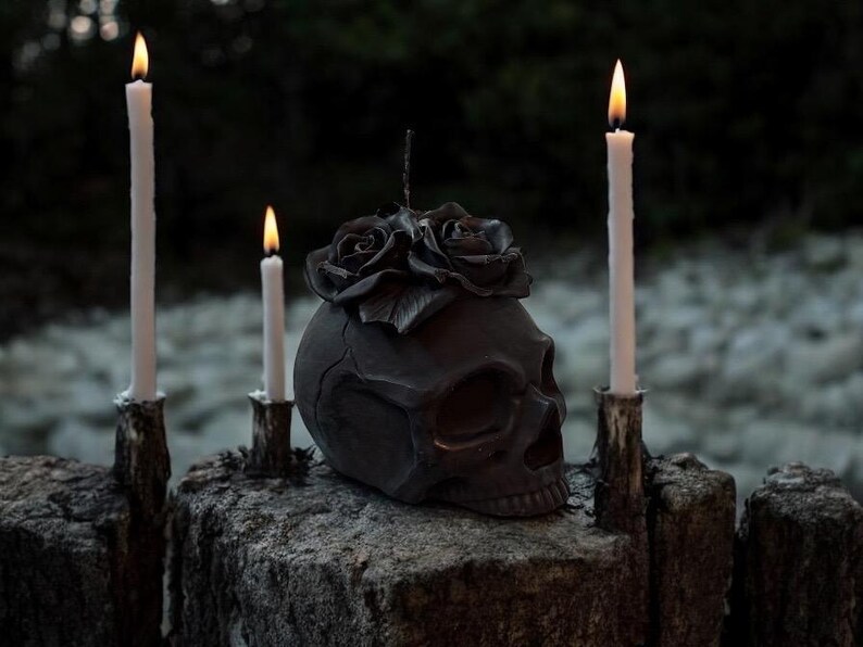 May include: A dark gray skull-shaped candle holder with a floral design on top, flanked by three lit white candles. The candles are in rustic holders, creating a gothic or Halloween theme. The background is blurred, suggesting an outdoor setting.