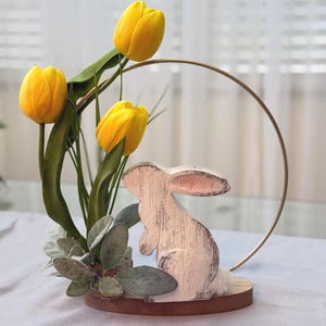 May include: A decorative Easter centerpiece featuring a wooden bunny with a distressed white finish, set on a round wooden base. The arrangement includes three bright yellow tulips, green leaves, and faux greenery, accented by a gold metal ring.