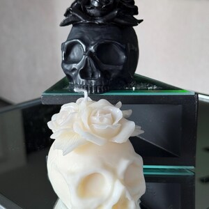 May include: Two skull-shaped candles. One is black with a black rose on top, the other is ivory with a white rose. The candles are on a reflective surface, creating a mirror image. The candles are decorative and gothic-themed.