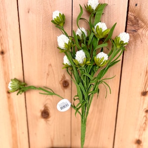 May include: A bunch of white faux flower stems with green leaves. The flowers have a fuzzy texture and are arranged in a bouquet. The stems are long and slender, and the leaves are small and delicate. The flowers are arranged in a vase, and the background is a wooden wall.