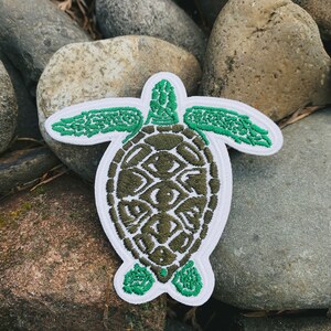 Puede incluir: Parche bordado con un diseño de tortuga marina verde y un borde blanco. El caparazón de la tortuga es verde oliva, y la cabeza y las aletas son de un verde vibrante. El parche se coloca sobre un fondo de rocas grises y marrones.