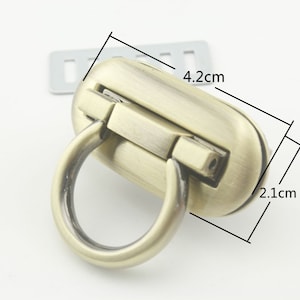 May include: Antique brass oval purse clasp with a ring handle. The clasp measures 4.2 cm long and 2.1 cm wide. A small, rectangular metal piece is visible in the background. This hardware is suitable for bags and purses.
