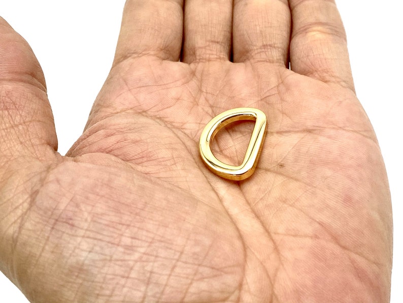 May include: A gold-colored metal D-ring hardware. The ring is smooth and has a polished finish.