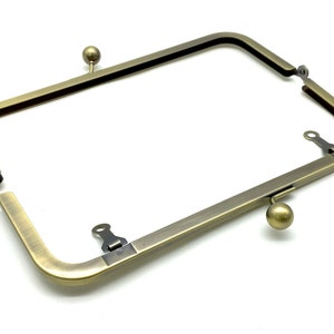 May include: Antique brass-toned metal purse frame with two clasps and two round knobs. The frame is open and ready to be used for a purse or clutch.