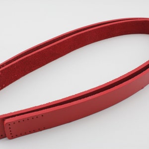 Pode incluir: Duas tiras de couro vermelho com costuras e furos para prender a uma bolsa ou outro projeto.