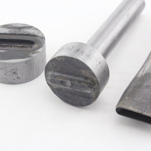 Op de afbeelding: Drie metalen lederen ponsgereedschappen. De gereedschappen zijn zilverkleurig en hebben verschillende rechthoekige gaten. De gereedschappen bevinden zich op een wit oppervlak.