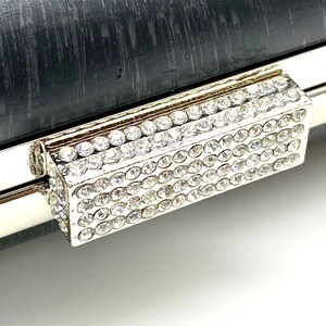 Pu&ograve; includere: Una pochette argentata con una chiusura rettangolare di strass. La chiusura &egrave; realizzata con piccoli strass trasparenti incastonati in una cornice argentata.