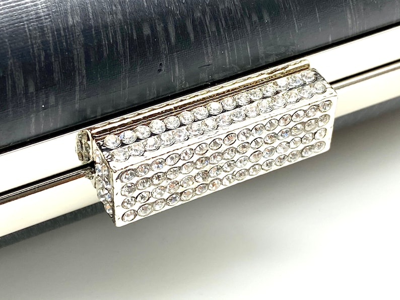 Pu&ograve; includere: Una pochette argentata con una chiusura rettangolare di strass. La chiusura &egrave; realizzata con piccoli strass trasparenti incastonati in una cornice argentata.