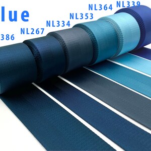 May include: A collection of blue nylon webbing rolls arranged in a row, featuring various shades from dark navy to light sky blue. Each roll is labeled with codes NL386, NL267, NL334, NL353, NL364, and NL339. The fabric has a distinct woven texture.