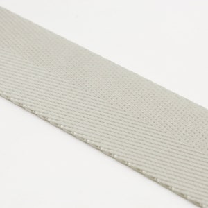 May include: A close-up of a light gray woven webbing strap. The strap has a herringbone pattern and a slightly textured surface.