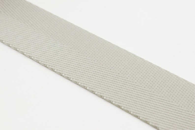 May include: A close-up of a light gray woven webbing strap. The strap has a herringbone pattern and a slightly textured surface.