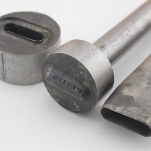 Puede incluir: Tres herramientas de punzonado de metal con aberturas ovaladas y rectangulares de diferentes tamaños. Las herramientas son de metal plateado y tienen un aspecto desgastado.