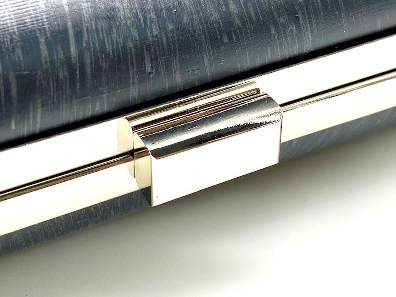 Pu&ograve; includere: Primo piano di una chiusura in metallo argentato su una pochette grigia. La chiusura ha una forma rettangolare e una finitura lucida.