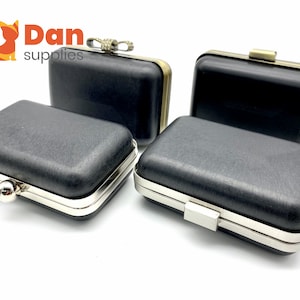 May include: Four black rectangular clutch bags with silver and gold metal frames. The bags have a textured fabric exterior and are closed with a metal clasp.