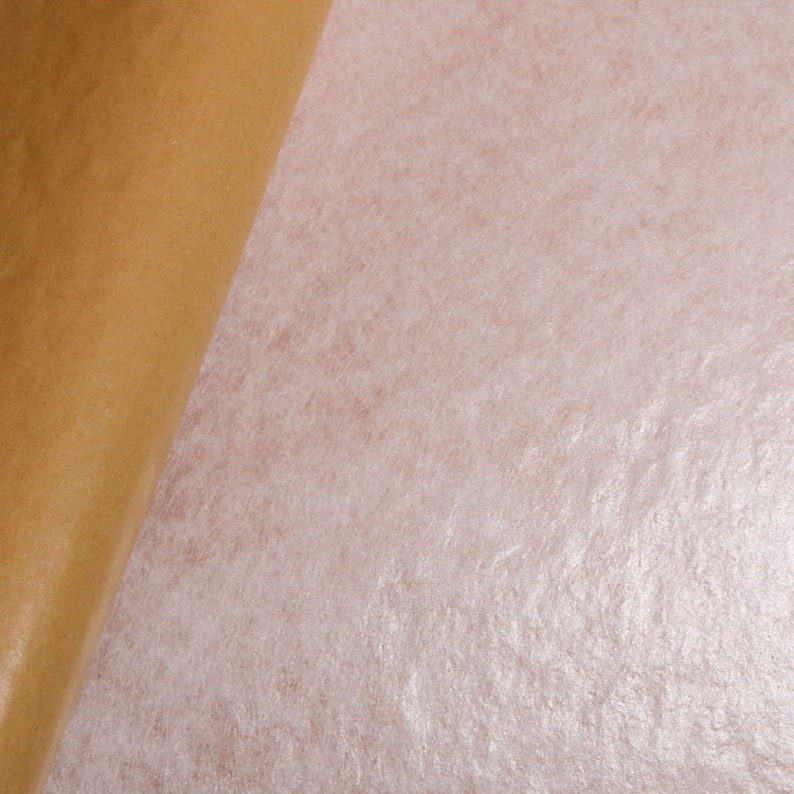May include: A close-up of a sheet of white paper with a subtle shimmery texture. The paper is being peeled back from a brown paper backing.