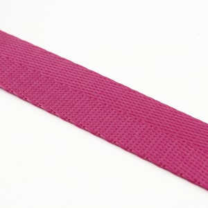 May include: A close-up of a length of magenta-colored webbing material. The webbing has a tightly woven, textured surface. The material is angled across the frame against a white background. This durable webbing is suitable for various crafting and DIY projects.