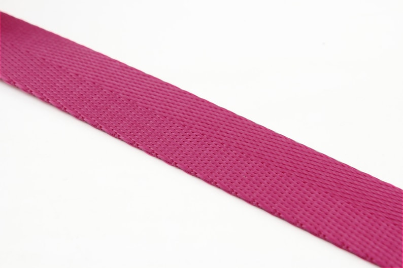 May include: A close-up of a length of magenta-colored webbing material. The webbing has a tightly woven, textured surface. The material is angled across the frame against a white background. This durable webbing is suitable for various crafting and DIY projects.