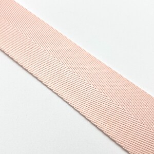 May include: Close-up of a blush pink herringbone weave ribbon. The ribbon is angled across a white background. The weave pattern creates a subtle chevron design. The ribbon is likely used for crafting, sewing, or decorative purposes.
