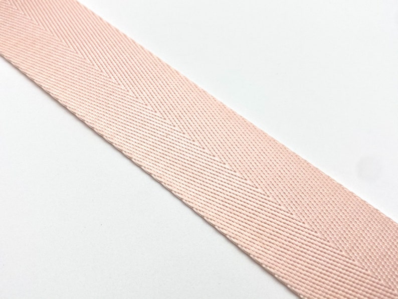 May include: Close-up of a blush pink herringbone weave ribbon. The ribbon is angled across a white background. The weave pattern creates a subtle chevron design. The ribbon is likely used for crafting, sewing, or decorative purposes.