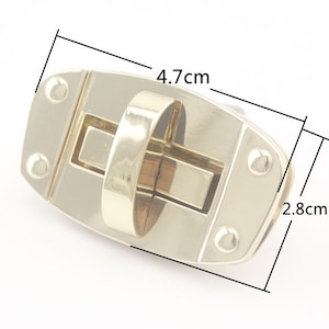 Könnte beinhalten: Goldfarbener Metallverschluss mit rechteckiger Öffnung und einem Ringgriff. Der Verschluss misst 4,7 cm x 2,8 cm.
