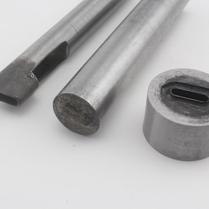 Op de afbeelding: Drie metalen gereedschappen voor het ponsen van gaten in leer. De gereedschappen zijn gemaakt van zilverkleurig metaal en hebben een afgeronde vorm. Het grootste gereedschap heeft een ronde basis en een gat in het midden. De andere twee gereedschappen zijn lang en dun met een gat aan het ene uiteinde.