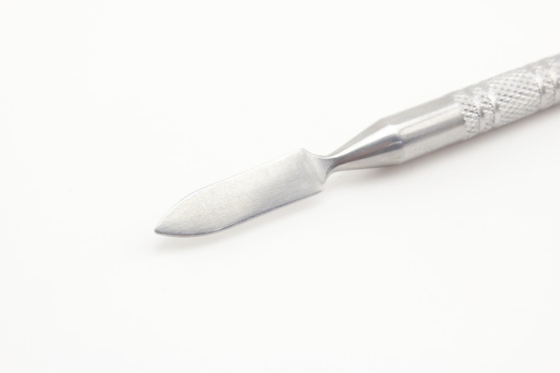 Op de afbeelding: Een zilveren metalen gereedschap met een puntige punt en een getextureerde handgreep. Het gereedschap wordt gebruikt voor schrapen en verspreiden.