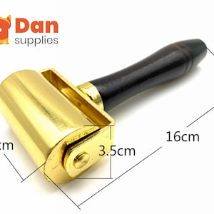May include: A gold-colored metal roller with a dark wooden handle, measuring 6 cm wide, 3.5 cm high, and 16 cm long. The roller is designed for crafts and leatherwork. The logo "Dan supplies" is in the top left corner.
