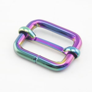 Op de afbeelding: Een regenboogkleurige metalen gesp voor een riem of band. De gesp is rechthoekig met afgeronde hoeken en heeft een gladde afwerking.
