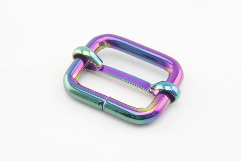 Op de afbeelding: Een regenboogkleurige metalen gesp voor een riem of band. De gesp is rechthoekig met afgeronde hoeken en heeft een gladde afwerking.