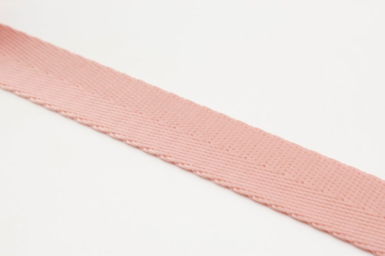 May include: A close-up of a length of pink woven fabric, likely a ribbon or strap, against a white background. The fabric has a diagonal weave pattern and a slightly textured surface. The color is a soft, muted pink.