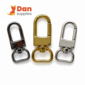 Könnte beinhalten: Drei drehbare Karabinerverschlüsse aus Metall in den Farben Gunmetal, Gold und Silber. Jeder Verschluss verfügt über einen rechteckigen Federbügel, eine drehbare Basis und einen D-Ring. Das Logo Dan supplies ist oben links zu sehen.