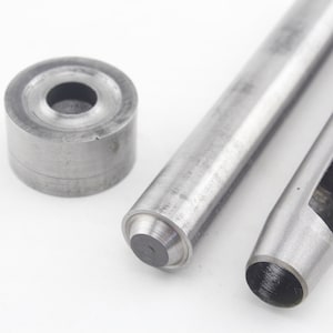Op de afbeelding: Een set metalen gereedschappen voor leerbewerking of vergelijkbare projecten. De set bevat een cilindrische pons, een massieve metalen pons en twee holle ponsen. De gereedschappen zijn gemaakt van staal en zijn ontworpen voor het maken van gaten in diverse materialen.