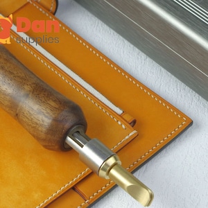 Könnte beinhalten: Nahaufnahme eines Lederbearbeitungswerkzeugs mit einem Holzgriff und einer Metallspitze. Das Werkzeug liegt auf einem Stück braunem Leder mit weißen Nähten. Der Text "Dan supplies" ist im Hintergrund sichtbar.
