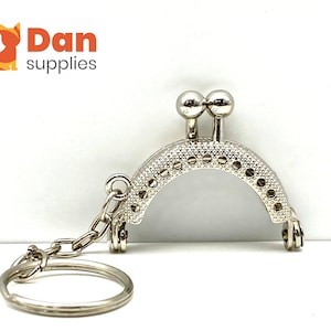 May include: Silver-toned purse frame with a key ring attached. The frame has a decorative pattern and two round metal accents at the top.
