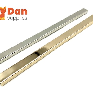 May include: Two metal trim strips, one silver and one gold, are shown on a white background. The strips are rectangular in shape and have a smooth, polished finish.