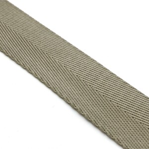 May include: A close-up of a beige herringbone webbing strap. The strap is made of a woven fabric with a textured surface.