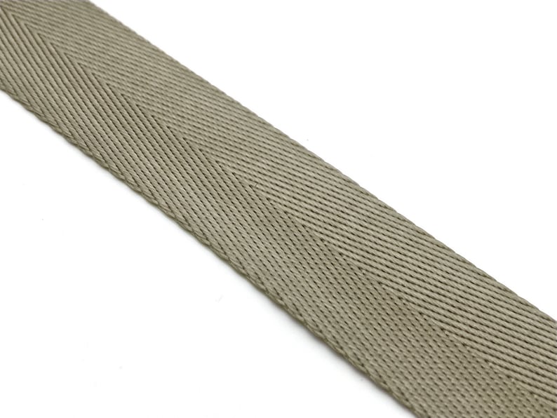 May include: A close-up of a beige herringbone webbing strap. The strap is made of a woven fabric with a textured surface.