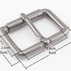 Może przedstawiać: Srebrna metalowa klamra o kwadratowym kształcie i zaokrąglonych krawędziach. Klamra ma wymiary 3,5 cm x 5 cm.