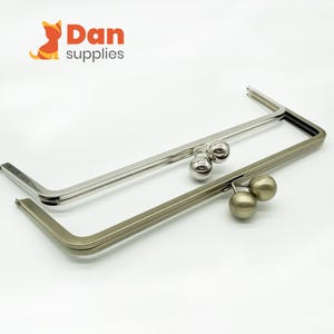 Puede incluir: Dos marcos de metal para monederos en tonos plateados y bronce. Cada marco tiene forma rectangular con esquinas redondeadas y un cierre de bola. El logotipo "Dan supplies" está en la esquina superior izquierda.